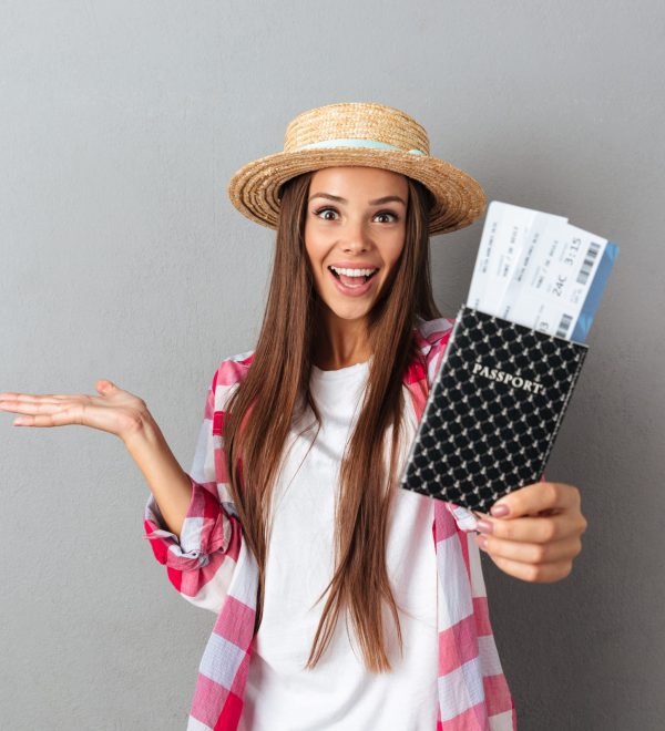 Close up portrait of a smiling happy woman traveller in straw hat showing passport with plane tickets looking at camera, over gray background