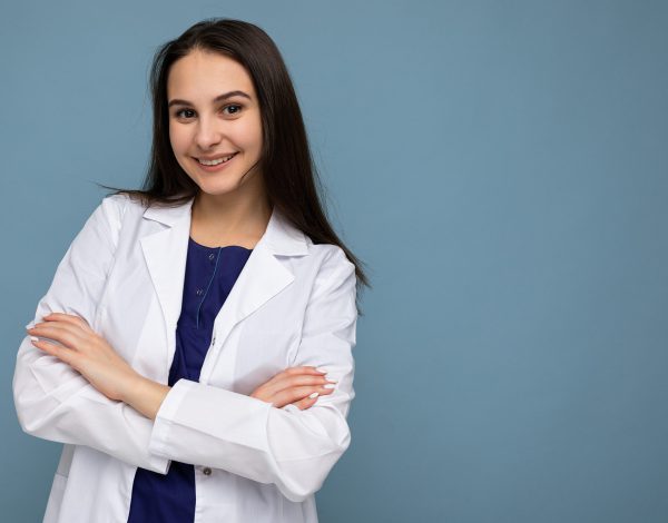 Photo portrait of young pretty beautiful positive smiling brunette woman with sincere emotions wearing white medical coat isolated over blue background with copy space and holding crossed arms.