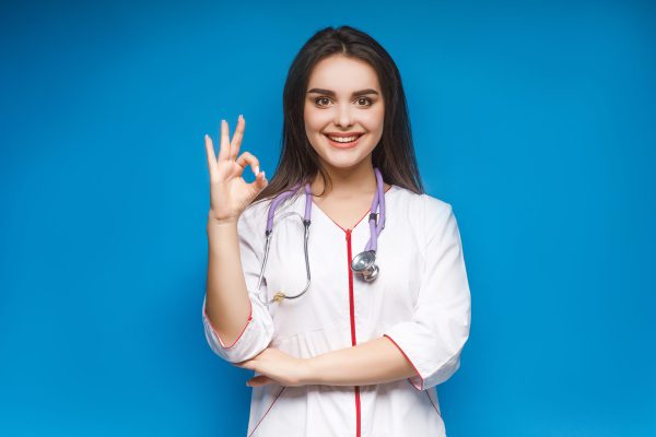 Photo of young female doctor make okaysign, over blue background.