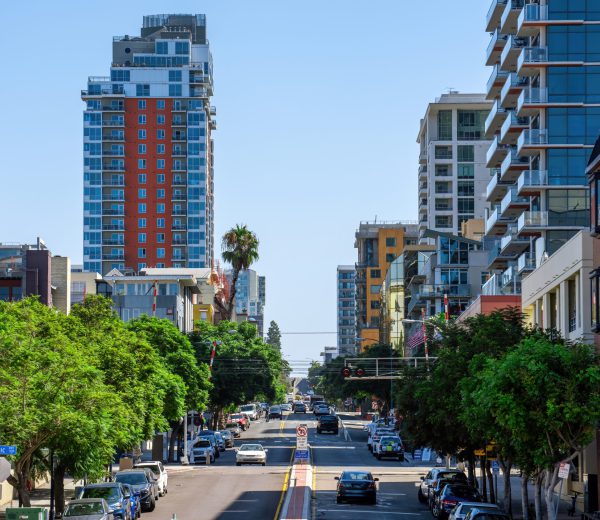 Road with multiple parked cars, green trees, modern buildings along it, sunny day in San Diego, USA