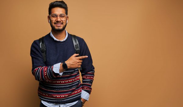 Smiling happy indian student with backpack pointing his finger on background.