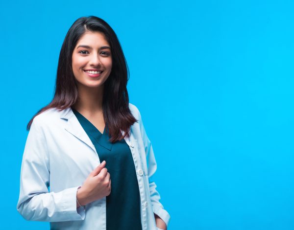 Medical concept of Indian beautiful female doctor in white coat with stethoscope, waist up. Medical student. Woman hospital worker looking at camera and smiling, studio, blue background