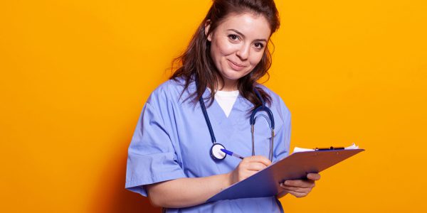 Cheerful nurse signing documents on clipboard, looking at camera. Medical assistant wearing uniform, writing on notebook papers and files smiling. Woman with stethoscope and textbook