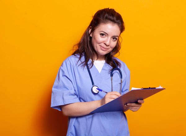Cheerful nurse signing documents on clipboard, looking at camera. Medical assistant wearing uniform, writing on notebook papers and files smiling. Woman with stethoscope and textbook