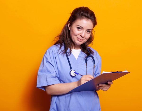 Cheerful nurse signing documents on clipboard, looking at camera. Medical assistant wearing uniform, writing on notebook papers and files smiling. Woman with stethoscope and textbook