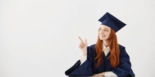 Young female university graduate in academic cap sitting at table smiling pointing left. Future lawyer or engineer showing an idea. White background. Copy space.